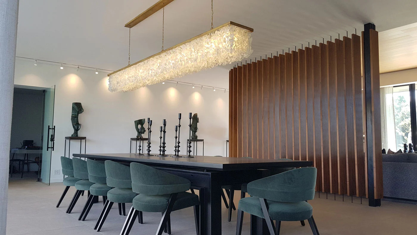 A modern farmhouse style home with picture windows at night with warm lighting inside where a kitchen and dining room are visible though the windows.